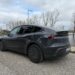 Tesla Model Y elétrico preto estacionado à beira de um lago, com árvores ao fundo, em um dia nublado.